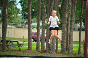 Young happy child girl playing alone, flying high on swings on summer weekend sunny day. Safety and recreation on playground concept