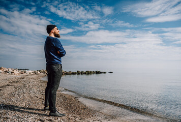 cooler junger Mann mit Bart, Sonnenbrille und Mütze steht bei blauem Himmel am Strand und sieht in die Ferne.