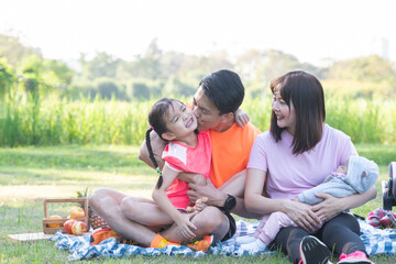 Happy Asian family in sportswear having a picnic at park, father kissing and hugging cute little daughter, mother smiling and holding newborn baby in arms, sitting on grass together. Family bonding