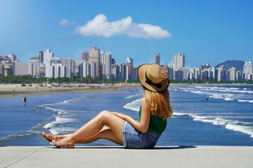Beautiful young woman holding hat sitting on ground with metropolis skyline and waving ocean on...