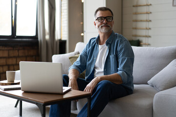 Senior man in casual clothing using laptop and smiling while sitting on the sofa, working from home.