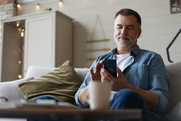 Happy smiling senior man using smartphone device while sitting on sofa at home - 582816358