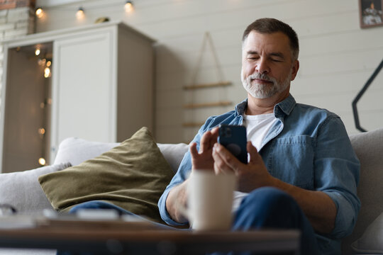 Happy smiling senior man using smartphone device while sitting on sofa at home