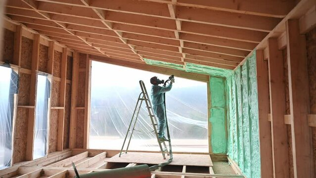 Male builder insulating wooden frame house. Man worker spraying polyurethane foam inside of future cottage, using plural component gun. Construction and insulation concept.