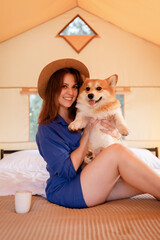 Happy woman in hat with Welsh Corgi Pembroke dog relaxing in glamping on summer day. Luxury camping tent for outdoor recreation and recreation. Lifestyle concept