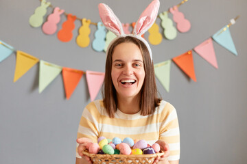 Portrait of attractive happy brown haired woman wearing rabbit ears holding Easter eggs, looking at camera with happy face, painting eggs for holiday, isolated on gray decorated background