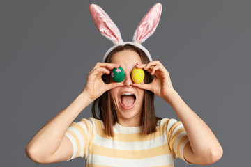 Happy Easter. Indoor shot of surprised funny crazy woman wearing rabbit ears covering her eyes with colorful Easter eggs isolated on gray background, keeps mouth open.