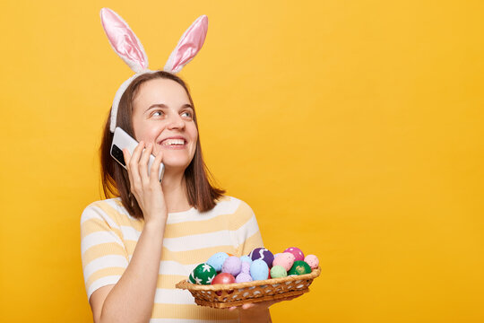 Image of attractive glad woman wearing rabbit ears holding Easter eggs and talking via mobile phone isolated on yellow background, advertisement area, copy space for promotion