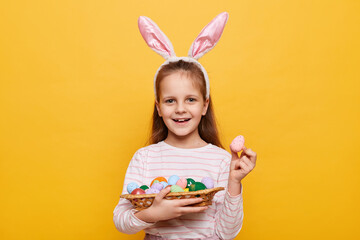 Obraz na płótnie Canvas Horizontal shot of cute optimistic little girl wearing rabbit ears holding Easter eggs in wicker basket, showing testicle painted by herself, isolated on yellow background