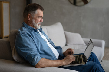 Senior man in casual clothing using laptop and smiling while sitting on the sofa, working from home.