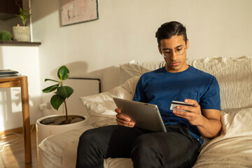 Attractive Hispanic man with glasses wearing blue shirt in the apartment using tablet and looking...