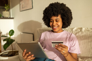 Obraz na płótnie Canvas Smiling girl with afro hairstyle wearing pink shirt and jeans in the apartment on the tablet holding a credit card in pink shirt and jeans