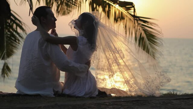 Newlyweds couple enjoys atmosphere of sunset against sun-path on ocean. Couple hugs under palm branch on beach after wedding ceremony