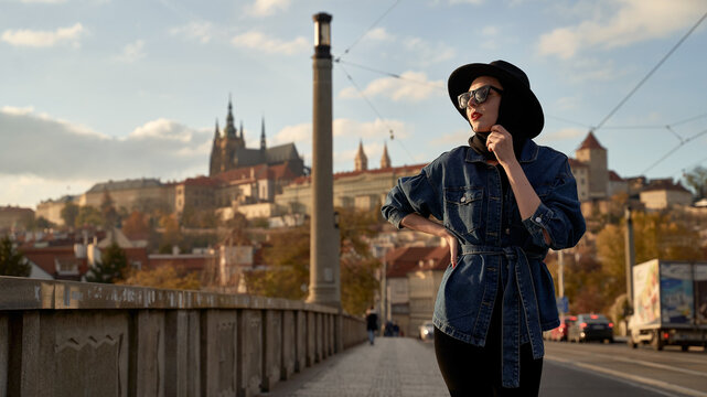 Stylish beautiful young woman earing black hat in Prague on a bridge with Prague Castle or Prazsky Hrad on background. Elegant retro lady fine art portrait.