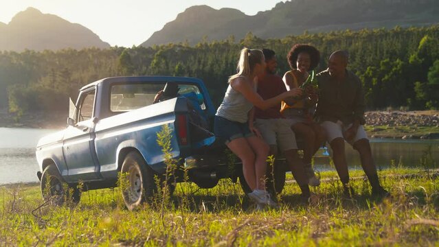 Friends with backpacks sitting on tailgate of pick up truck on road trip in countryside drinking beer and relaxing  - shot in slow motion