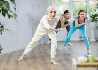 Obraz premium Cheerful sporty senior woman training movements of rhythmic latin zumba with group in fitness studio. Concept of active lifestyle of aged adults