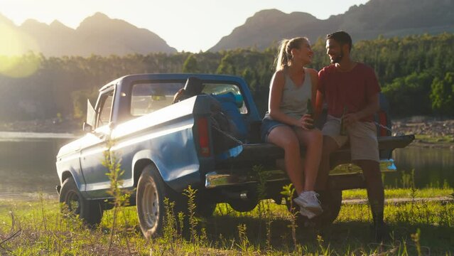 Couple with backpacks sitting on tailgate of pick up truck on road trip in countryside drinking beer and doing cheers  - shot in slow motion