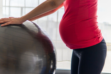 Closeup on a pregnant woman in a vibrant sports shirt leaning on an exercise ball and stretching against a window. Pregnancy, maternity, motherhood, sport, yoga, and health concept. Bright background.