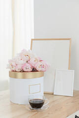 Vertical frame and gift box mockup on a wooden table in the kitchen. Glass jug with a bouquet of pink peonies and a cup of black coffee. Scandinavian style interior.