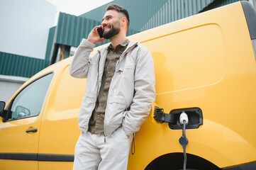 bearded caucasian man standing near an electric car that is charging and making time adjustments on a smartphone.