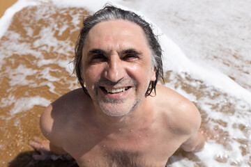 Fototapeta na wymiar Adult man sitting on yellow sand in a foam wave, Greece, Crete, Chania
