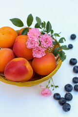  Still life with ripe apricots and blueberries, fruits with pink roses in a yellow basket on a white background, closeup, summer wallpaper.