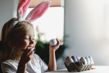 Cute little smiling girl wearing bunny ears is preparing painted Easter eggs for Easter sitting at the kitchen table. Happy Easter. Adorable child celebrate Easter at home. 