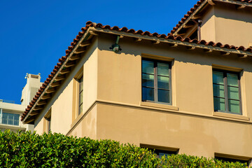 Beige stucco cement building with dark roof tiles in midday sun with front yard shrubs and copy space blue sky