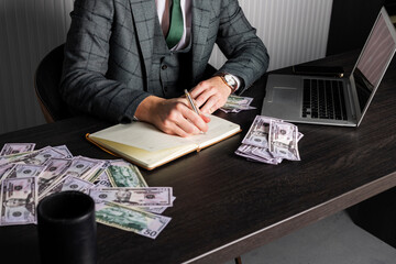 Close-up of a young serious man in a gray formal suit sits at his desk surrounded with cash and takes a note in his notebook. A businessman at work.