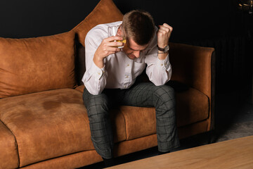 A young upset man in a white shirt sits on a brown couch with a glass of whiskey.