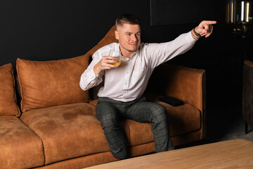 A young man in a white shirt sitting on a couch pointing at TV with glass of whiskey in other hand.