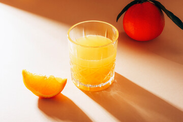 A glass of orange juice and slice of orange fruit on beige table with long hard shadows. Healthy raw vegan food, diet concept. Closeup