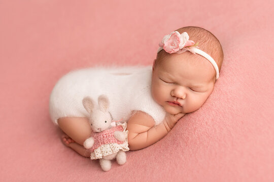 Top view of a newborn baby girl sleeping in a white overalls, with a white bandage on her head. With a knitted white rabbit on a pink background. Beautiful portrait of a newborn baby 7 days, one week.