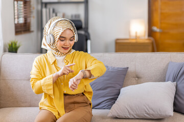 Young beautiful brunette muslim woman wearing arab hijab on sofa at home, Checking the time on wrist watch, relaxed and confident