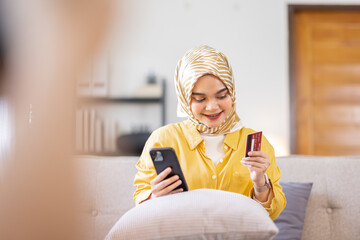 Easy payments. Smiling arabic woman or muslim woman in hijab in headscarf using phone and credit card at home, paying for utilities online, free space