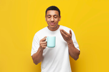young african american guy in white t-shirt holds cup of coffee and smells the aroma on yellow isolated background