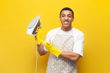 African American guy in apron and gloves holds iron on yellow isolated background, male housekeeper irons clothes - obrazy, fototapety, plakaty