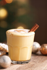 Tasty eggnog with cinnamon, cookies and baubles on wooden table against blurred festive lights