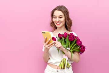 young attractive woman in white festive clothes holding bouquet of pink tulips and using smartphone