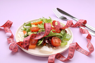 Plate with fresh vegetable salad and measuring tape on violet background, closeup. Healthy diet concept