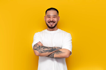 adult tattooed asian man in white t-shirt stands with his arms crossed on yellow isolated background and smiles