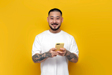 adult tattooed asian man in white t-shirt uses smartphone on yellow isolated background and smiles