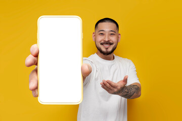adult tattooed asian man in white t-shirt shows empty smartphone screen mock-up on yellow isolated...