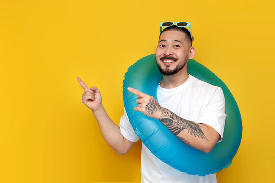 tattooed male asian tourist in white t-shirt and sunglasses holding inflatable swim ring and pointing to the copy space