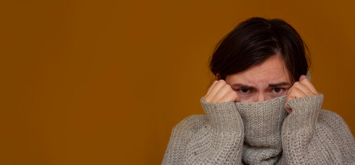 Horizontal banner. Young frightened mad woman with dark hair covers her face with wool sweater collar in fear on mustard background. Concept of childlessness, childfree, domestic violence, protection