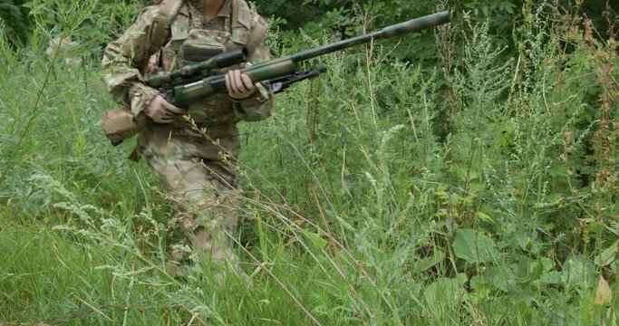 female soldier is walking in green forest.she takes aim from a assault rifle while sitting in shotting position slow motion, girl is running to take a position