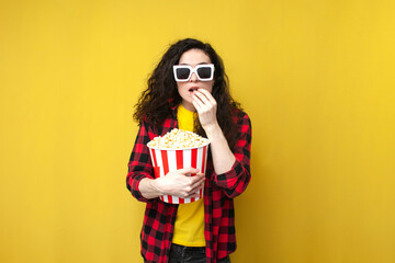 curly girl in 3d glasses watches movie and eats popcorn on yellow isolated background, brunette woman is cinema viewer