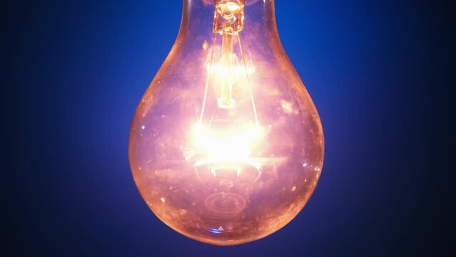 Incandescent lamp lights up and flickers on blue background. A light bulb turned on and off close-up. Warm flashing filament. Tungsten bulb light and cozy shine of vintage light. Idea, copy space. 4K
