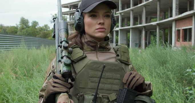 Young serious confident woman in military uniform armed with rifle looking sideway, girl with gun on her shoulder pretecting area, territory ca,mera motion security ruined biuilding in the background