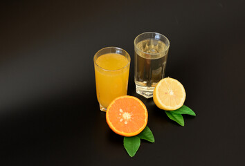 Two tall glass glasses with orange and lemon juice, next to pieces of ripe citrus fruits with green leaves on a black background.